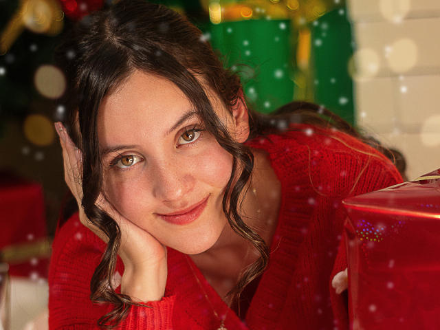 ChloeDulce, con i capelli castano ondulati, un maglione rosso e un sorriso carino, appoggia la testa sulla mano, guardando la fotocamera, circondata da regali di Natale e luci sfocate, mostrando leggermente il seno..