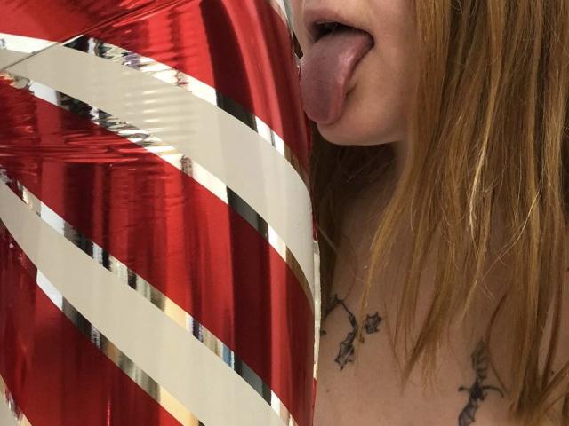 Rubbi with tongue out, red and white striped foil covering her tits, has tattoos on her chest, and is a fair-skinned redhead.