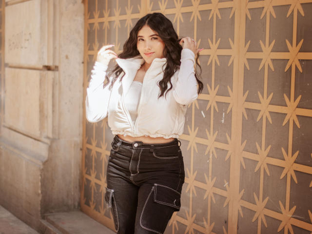 Veronica, avec ses cheveux noirs ondulés, son blouson blanc gonflé et son jean noir, se tient devant un mur à motifs, tenant le col de son blouson, souriant légèrement, et montrant son petit postérieur..