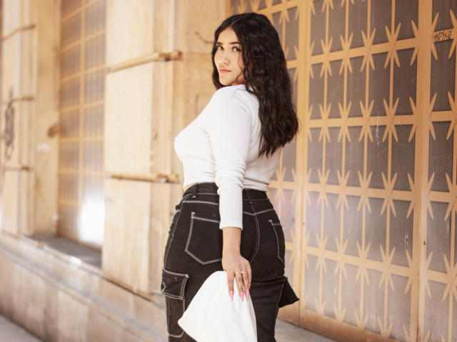 VeronicaWins with curly black hair, white long-sleeve top, black high-waisted skirt, holding white cloth, standing with back to camera, looking over shoulder, showing her ass.