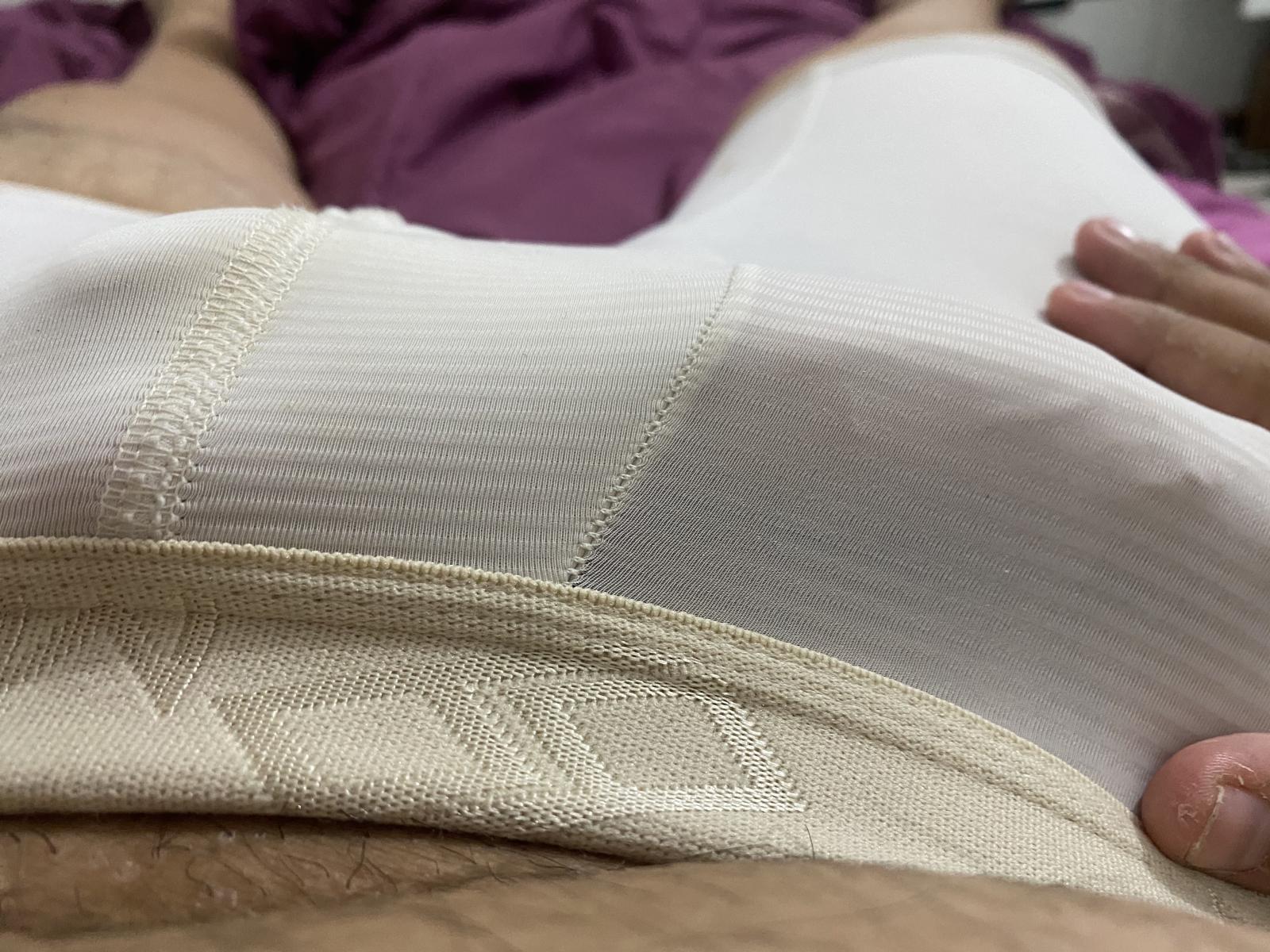 O macacão branco texturizado de JuanX69, que mostra seus seios e nádegas, com uma cobertura de renda bege, está sobre uma cama roxa. Uma mão com unhas rosa descansa em sua barriga..