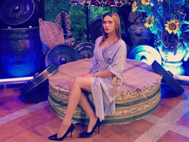 YanaHera, con cabello rubio, un vestido a rayas y tacones altos negros, sentada en una cama redonda con rayas verdes, mostrando sus piernas y mirando a la cámara. Los senos no son visibles..