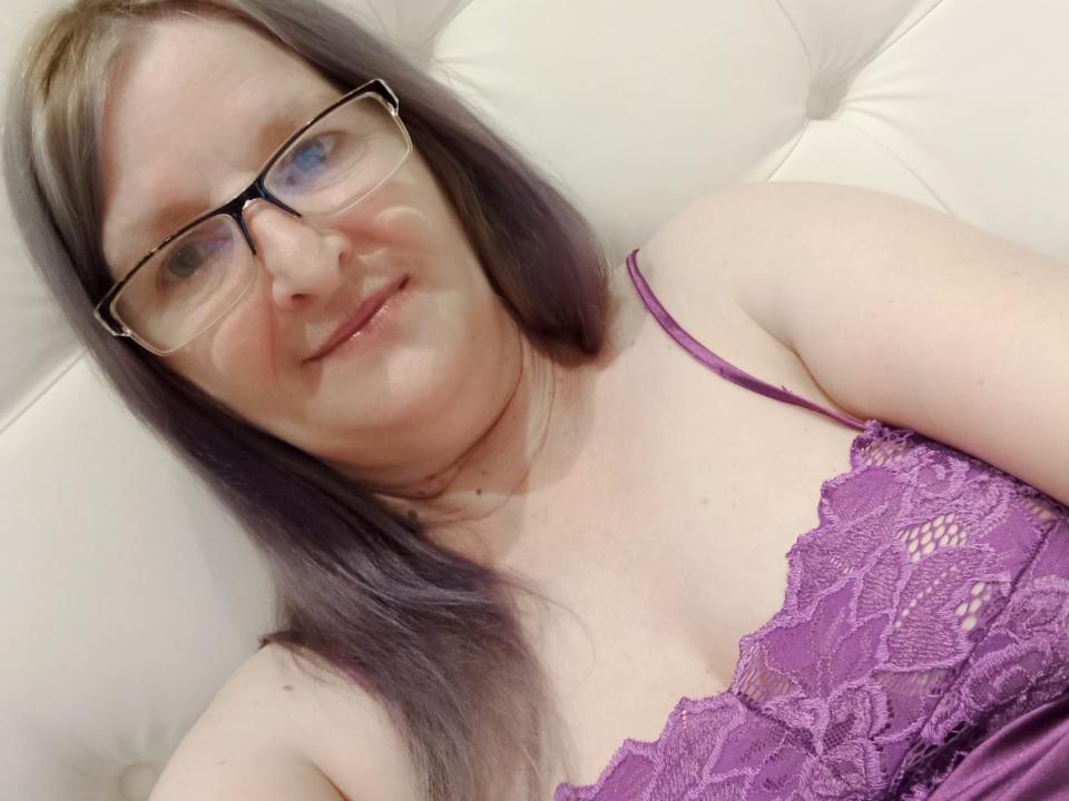FantasyLilla, a chubby woman with purple lace bra, glasses, and grayish-purple hair, smiling slightly. Her boobs are visible under the lace. She's lying on a white couch.