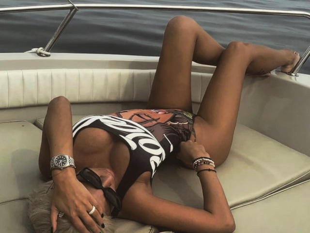 BeatriceGalaxy está acostada en un bote, usando un traje de baño blanco y negro, mostrando sus pechos, con las piernas separadas, y lleva gafas de sol, pulseras y un anillo, sobre un cojín..