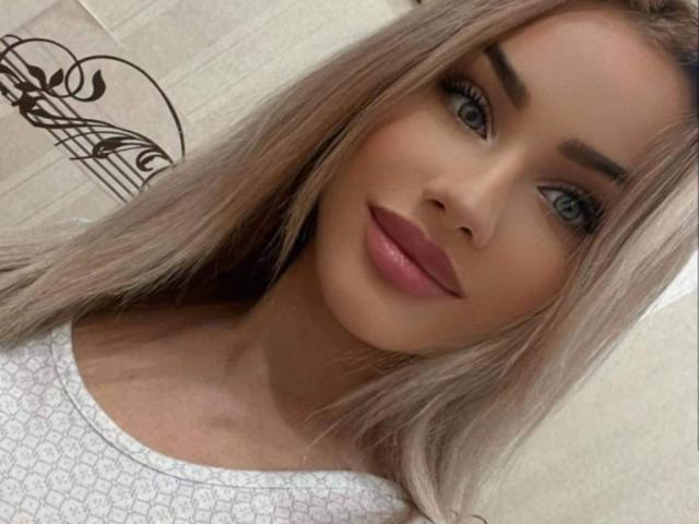 BeatriceGalaxy heeft blond haar, blauwe ogen en roze lippenstift. Ze draagt een witte top met een geruit halslijn. Haar achterkant is niet zichtbaar..