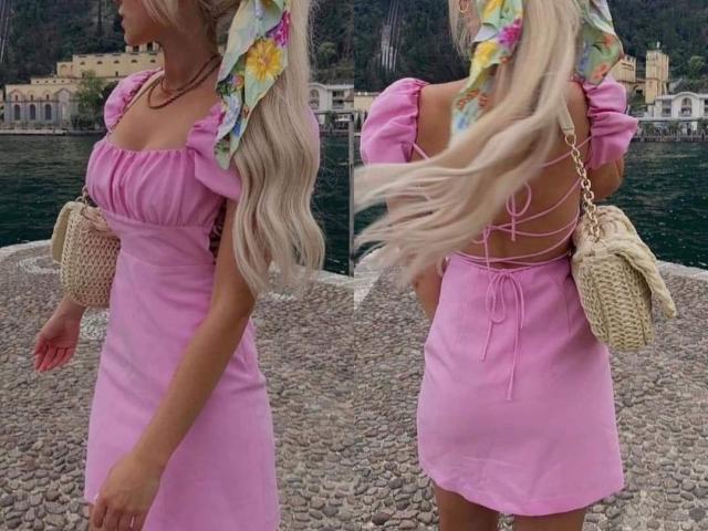 Beatrice, com longos cabelos loiros, vestindo um vestido rosa, lenço de cabeça floral, bolsa de vime e com o fechamento de renda nas costas visível. Seios levemente visíveis, vestido rosa justo, em pé perto da água..