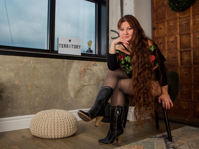 OVanessaHotly with long brown hair, wearing a black floral top, black sheer tights, and black cowboy boots, sitting on a chair, hand on cheek, next to a white pouf.