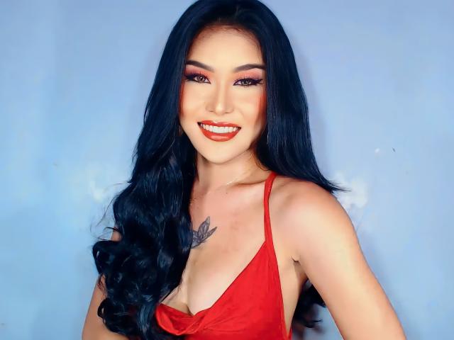 MayenLim with long black hair, red dress, and cleavage tattoo, smiling with red lipstick and bold eye makeup. Boobs, red dress, and tattoo visible. Smiling, sexy, and attractive.