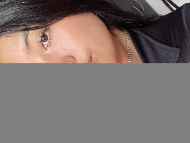 Elizaa, com olhos castanhos brilhantes, lábios rosados e brilhantes, cabelo preto e liso, um piercing no nariz, um colar de prata e uma jaqueta preta com zíper. Sua pele é lisa e ela está olhando para a câmera..