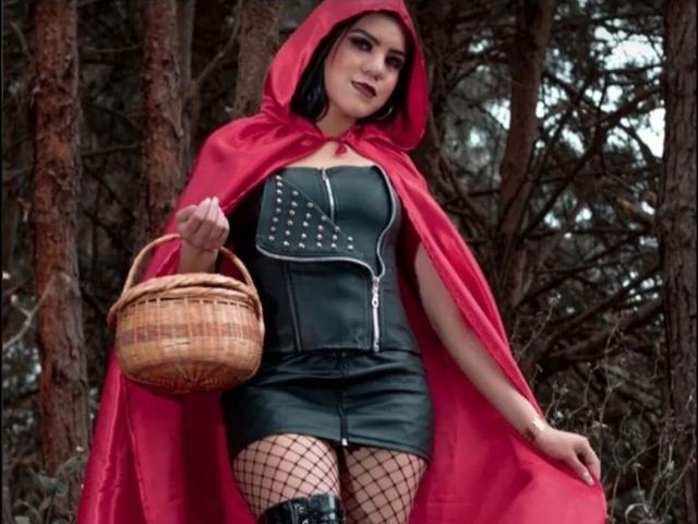 KarlyGreyKiss com capuz vermelho, corpete de couro preto e minissaia, segurando uma cesta de vime, usando meias de rede e um manto vermelho, em pé em uma floresta. Seios visíveis..