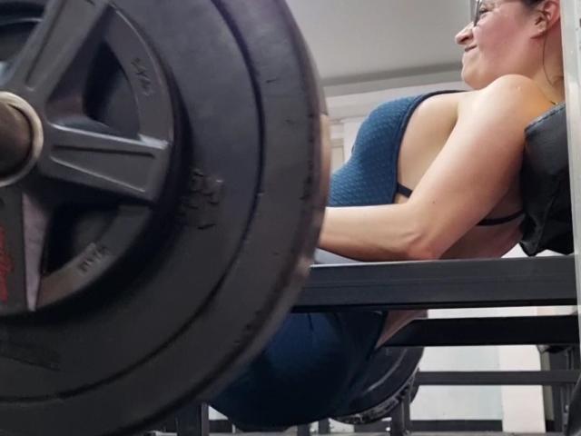 VaaleryStark's ass is raised off a black bench, gripping it with her hands, wearing a blue, textured sports bra, lifting a heavy black weight with a focused expression.