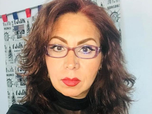 Catherine, eine Frau mit welligem, braunem Haar, lila Brille, rotem Lippenstift, einem schwarzen Rollkragenpullover, einem Nasenpiercing und einem ernsten Gesichtsausdruck. Brüste nicht sichtbar, Vulva nicht sichtbar, Brüste nicht sichtbar..