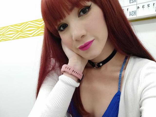 AkemixDiego heeft rood haar, draagt een blauwe beha, roze lippenstift, een zwarte choker, een witte cardigan en een roze horloge. Ze leunt haar hoofd op haar hand en glimlacht. Haar borsten zijn klein..
