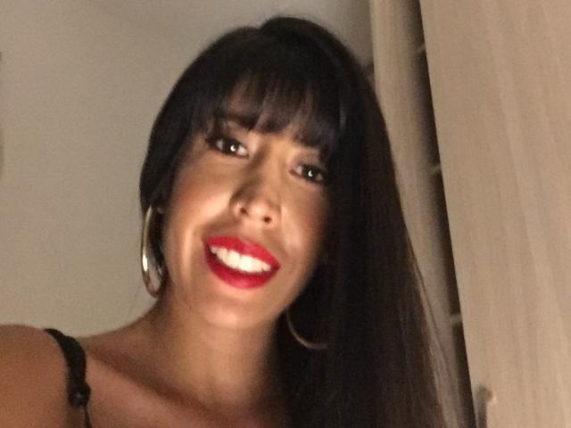 SeleneBrigth with dark straight hair, bangs, red lipstick, smiling, showing white teeth, wearing black strap, large hoop earrings, and has tan skin.