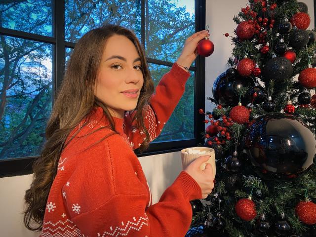 LuciaPerth, con lunghi capelli castani, un maglione natalizio rosso e fiocchi di neve bianchi, sta decorando un albero con ornamenti rossi e neri, tiene in mano una tazza bianca e sorride..