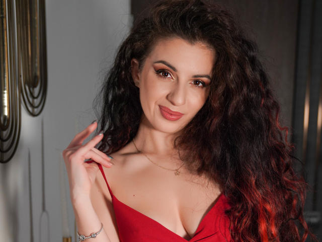 AnaAnais con capelli ricci e rossi scuri, vestito rosso, seno piccolo, sorridente, che tiene lo spallino del vestito, rossetto rosa, trucco leggero, braccialetto d'argento, collana, occhi marroni, illuminazione soffusa, sguardo sicuro..