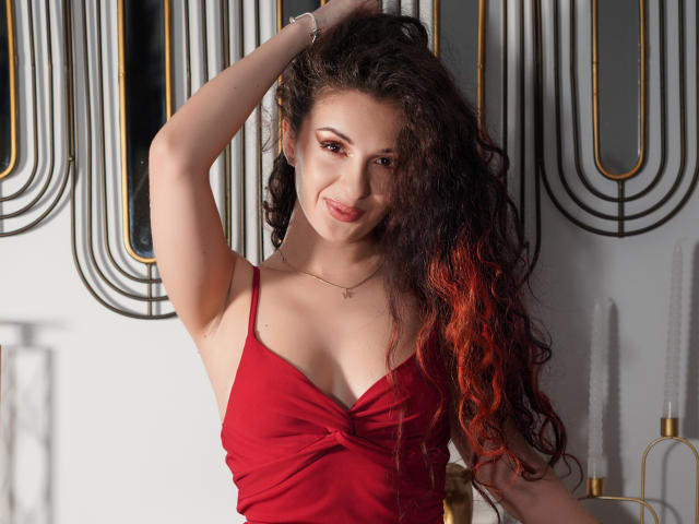 AnaAnais with curly dark red hair, wearing a red dress, has her arm raised, showing her tits, smiling, and looking at the camera. She's wearing a silver bracelet.