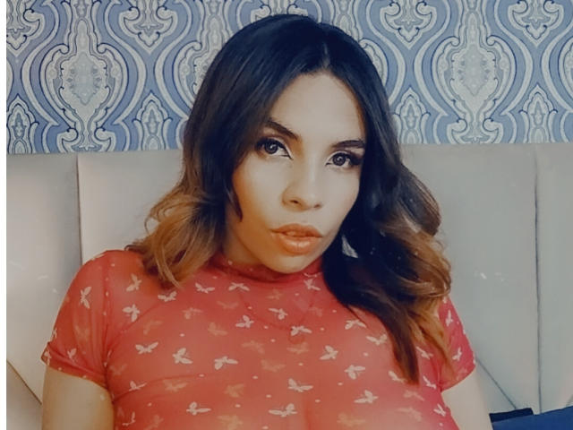 VioletaStone with long brown hair, wearing a red sheer top with butterfly patterns, showing her tits, sitting against a white cushioned background. She has a serious, slightly pouty expression.