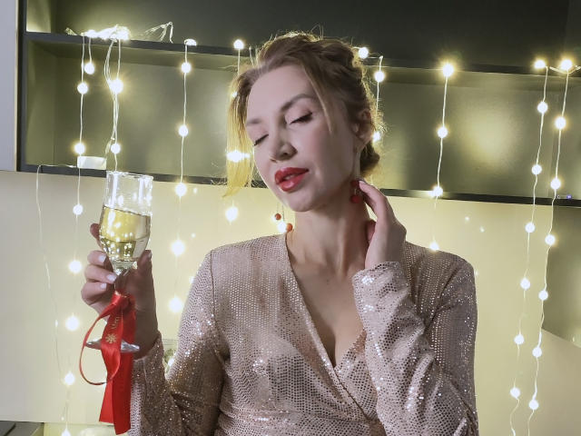 SimoniGrace, vêtue d'une robe rose scintillante, tenant une flûte de champagne, les yeux fermés, avec des boucles d'oreilles rouges, et des guirlandes lumineuses en arrière-plan. Elle a un joli visage et une légère moue..