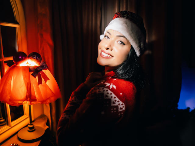 AnelisaReys with black hair, smiling, wearing a red Christmas sweater, white fur hat, and looking over her shoulder at a glowing orange lamp, showing her cute ass.