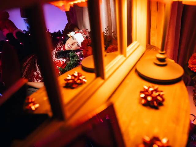 AnelisaReys décorée pour Noël, tenant une guirlande verte, portant une robe rouge et blanche, un ornement blanc pour la tête, regardant la caméra, à l'intérieur d'une structure en bois avec des lumières orange et des bougies..