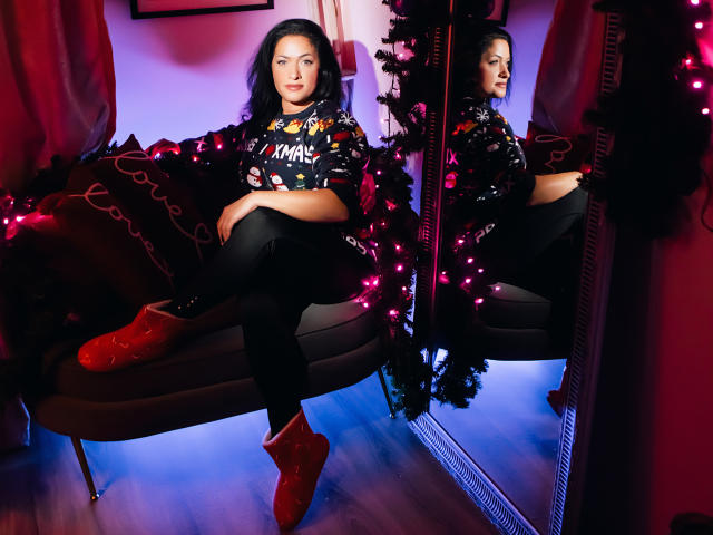 AnelisaReys with wavy black hair, red boots, black pants, and a black shirt with colorful prints, sitting on a gray couch, reflected in a mirror. Boobs, ass, mirror.