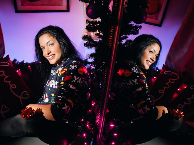 Anelisa Reys mit langen, schwarzen Haaren, lächelnd, trägt einen festlichen Pullover und sitzt vor einem Spiegel, der ihr Spiegelbild zeigt. Im Hintergrund steht ein Weihnachtsbaum mit roten Ornamenten und rosa Lichtern..