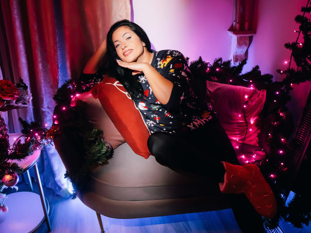AnelisaReys with dark hair, wearing a black floral shirt and black pants, sits on a pink-lit couch, hand on cheek, red pillow, Christmas lights, and green garland.