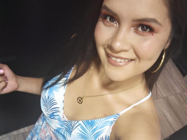 ConniRiccy with brown hair, smiling, wearing a blue and white tropical-patterned dress, gold hoop earrings, and a gold necklace with a black infinity symbol, showing a small red mark.