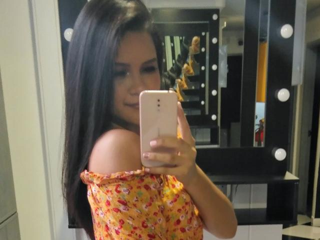 ConniRiccy with long dark hair, smiling, taking a mirror selfie in a dressing room. Wearing an off-shoulder, floral yellow dress. Holding a pink iPhone. Boobs visible.