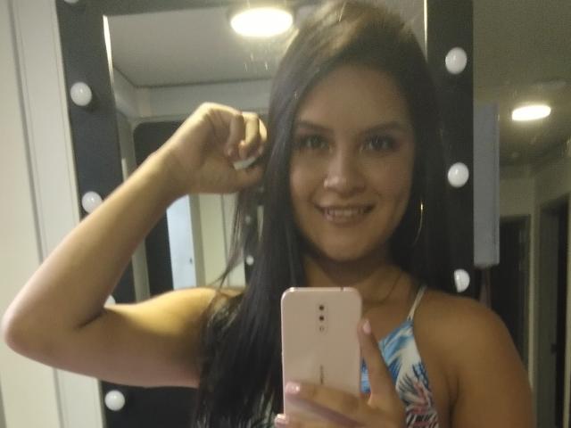 ConniRiccy, con cabello negro largo, sonriendo, sosteniendo un teléfono rosa, vistiendo una blusa con estampado azul y blanco, flexionando el brazo, espejo con bombillas en el fondo..