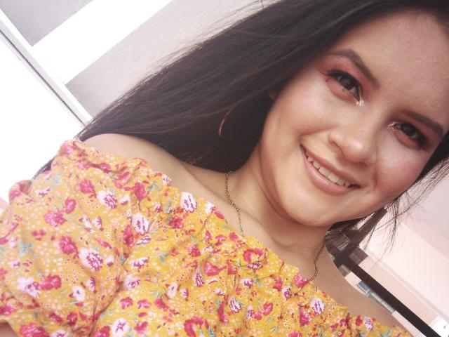 ConniRiccy, ragazza asiatica con lunghi capelli neri, che sorride, indossa una maglietta gialla a fiori con una spalla scoperta, ombretto rosa, una collana dorata, mostrando un viso carino e paffuto, un naso piccolo e labbra rosa leggermente dischiuse..