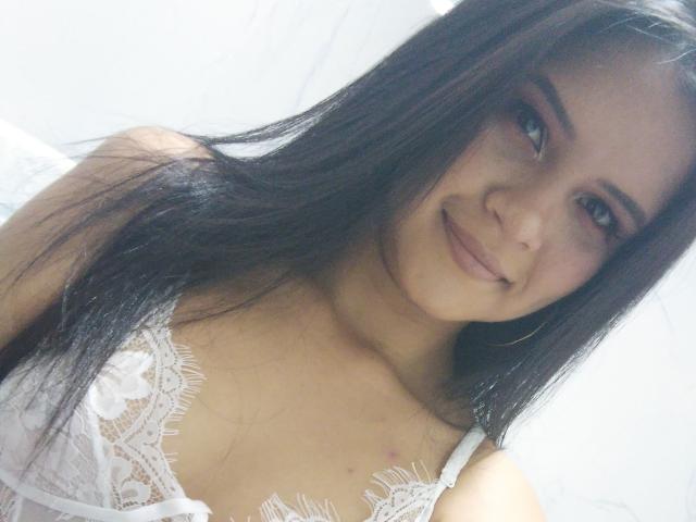ConniRiccy, com cabelos longos e pretos, usando um sutiã de renda branco, sorrindo para a câmera, mostrando seios pequenos, pele clara e maquiagem rosa nos olhos..