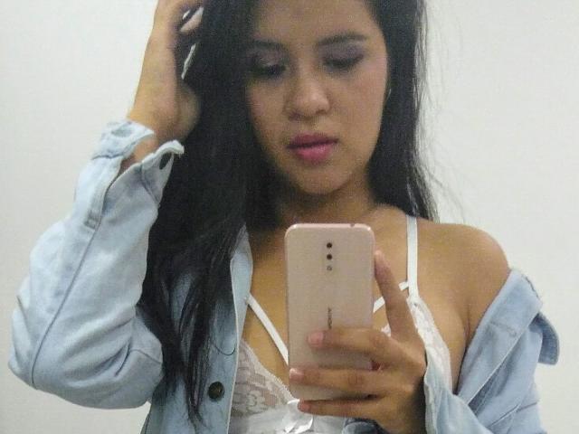 ConniRiccy with long black hair, pink lipstick, and white lace bra, holding a pink iPhone, wearing an open blue denim shirt, looking at her phone, hand in hair.