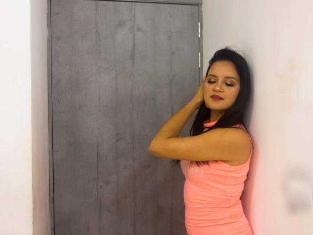 ConniRiccy, con lunghi capelli neri, rossetto rosso e canotta rosa, in piedi contro un muro, che tocca l'orecchio con la mano destra, con un'espressione leggermente annoiata..