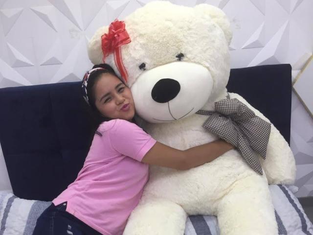 ConniRiccy abraza a un enorme oso de peluche blanco con un lazo rojo y una corbata de lunares. Lleva una camisa rosa y pantalones vaqueros azules, y está sonriendo a la cámara..