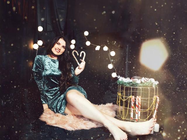 MilaLoona with long dark hair, green velvet dress, holding scissors, sitting on pink fur, legs extended, Christmas basket with candy canes, string lights, smiling, bare feet.