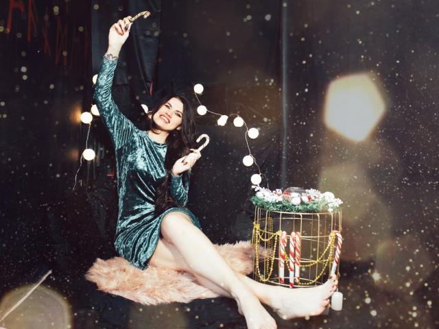 MilaLoona in a green velvet dress, sitting on a pink furry rug, decorating a Christmas tree with candy canes and string lights, holding a ornament, smiling.