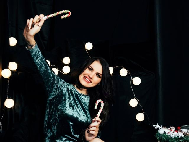 MilaLoona with long black hair, wearing a green velvet top, holding a candy cane in each hand, smiling, surrounded by glowing string lights.