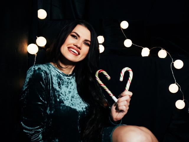 MilaLoona with long black hair, smiling, wearing a green velvet dress, holding two candy canes, standing against warm string lights, dark background. Boobs, ass not visible.