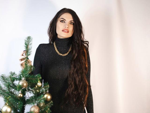 MilaLoona with long dark hair, wearing a black glitter turtleneck, gold chain necklace, standing by a decorated Christmas tree, holding a branch with gold ornaments.