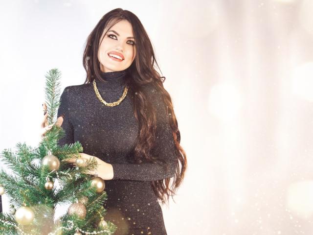 MilaLoona with long dark hair, wearing a black glittery turtleneck, gold necklace, smiling, holding a green Christmas tree with gold ornaments, standing in soft light.