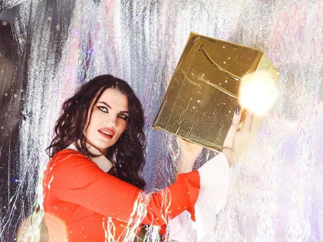 MilaLoona, con cabello negro ondulado, un vestido rojo y mangas blancas, sosteniendo un bolso dorado y brillante, sonriendo, sobre un fondo plateado y brillante. Sus senos son parcialmente visibles..