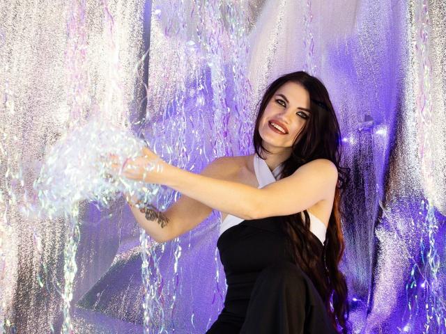 MilaLoona with long black hair, white halter top, black dress, smiling, splashing water with hands, showing tattoo on left forearm, in a glittery, purple-lit background.