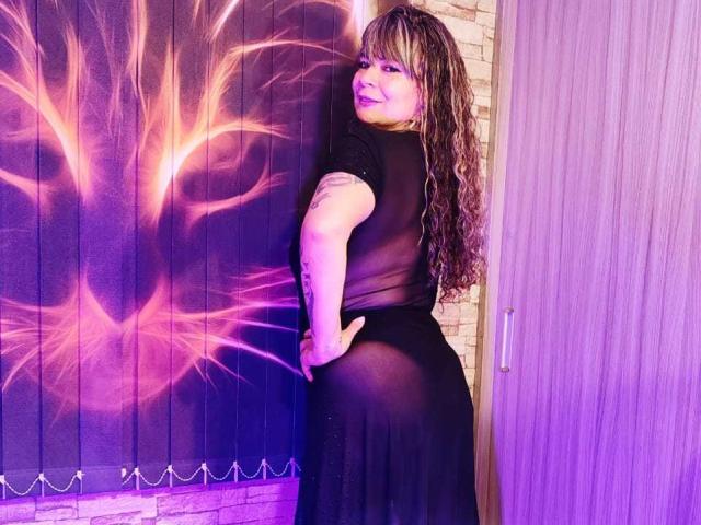 BrendaDollX in a sheer black dress, showing her ass, with long curly brown hair, smiling, hand on butt, tattoo on right arm, purple and pink light effect.