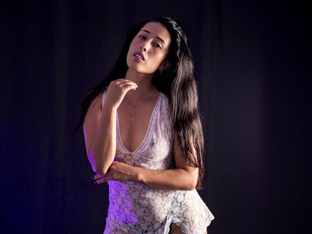 KatherinParmer with long black hair, wearing a sheer white lace top, exposing her tits, standing against a dark background, hand near her mouth, looking seductive.