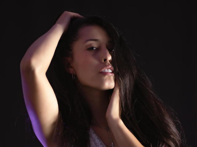 KatherinParmer, avec de longs cheveux foncés, les mains dans les cheveux, un léger sourire, une peau éclatante, de petites seins, un haut en dentelle blanche, un fond sombre, une lumière violette douce, sexy, attirante, confiante, buste..