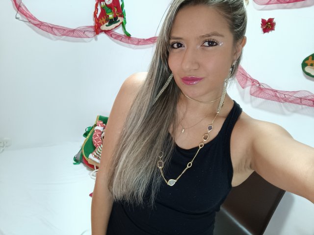 ValeriaHunter69 with long grayish-blonde hair, pink lipstick, black sleeveless top, gold necklace, earrings, and slight smile, holding phone for selfie, showing small tits and armpit.