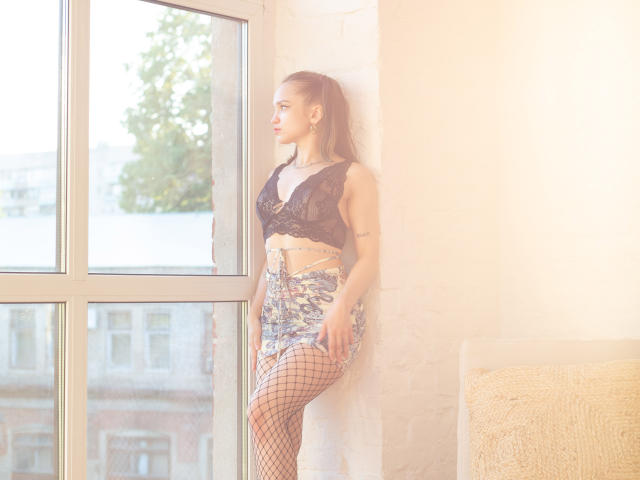 ProvocateurMur steht an einem Fenster in Fischernetzstrümpfen, einem Spitzen-BH, floralen Shorts und einem Pferdeschwanz, wobei das Sonnenlicht sie beleuchtet. Ihre Brüste sind klein, ihr Hintern ist kurvig..