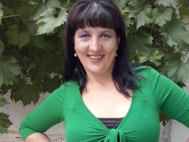 Ridemeraf with black hair, green top, and purple eyeshadow, smiling, showing tits, standing in front of grape leaves.